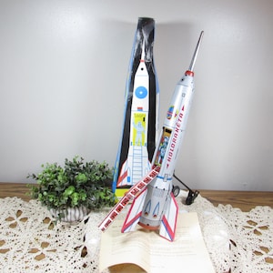 May include: A vintage tin toy rocket with a silver body, red and blue accents, and the text "HOLDRAKETA." The rocket is displayed with its original box, featuring a similar rocket design. A small plant and lace doily are also visible.