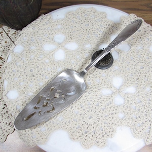 May include: A vintage silver cake server with a decorative handle and a slotted blade. The server rests on a white marble surface, atop a cream-colored, crocheted doily. The server is designed for cutting and serving cake.
