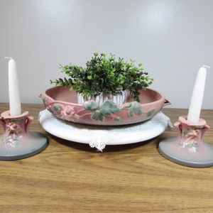 May include: A decorative arrangement featuring two pink and gray candle holders with white candles, a pink floral bowl, and a small green plant. The items are arranged on a white plate with lace trim.