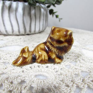 May include: A small, glossy ceramic figurine of a dog in a reclining position. The figurine features a mottled brown and tan glaze with a smooth, reflective finish, depicting the animal with its head turned and tail extended behind its body.