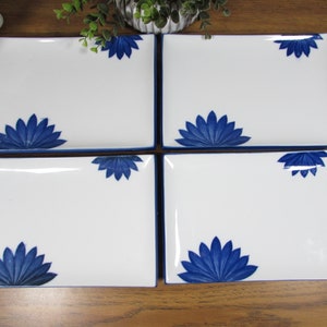 May include: Four white rectangular plates with a blue floral design in each corner. The plates have a blue border and are arranged on a wooden surface. The floral design is a stylized lotus or water lily.