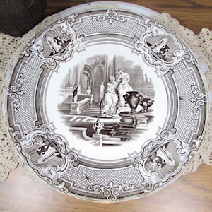 May include: A decorative plate with a brown and white design. The plate features a central scene of figures in an architectural setting, surrounded by ornate borders. The plate is placed on a cream-colored lace doily.