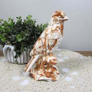 May include: A ceramic bird figurine with a brown and cream marbled pattern. The bird is perched on a brown base, and the figurine is set against a neutral background. The bird is a decorative item.