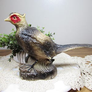 May include: A vintage ceramic pheasant figurine with a detailed design. The bird has a red face, a yellow beak, and brown and black feathers. It is perched on a brown base with green foliage in the background.
