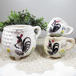 May include: White ceramic measuring cups and a pitcher with a rooster design. The pitcher has a textured surface and a handle. The cups are labeled "1/4 cup" and "1/8 cup". The rooster is black, white, and red.