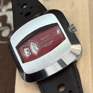 May include: A Sevan wristwatch with a silver-toned square case and a black perforated leather strap. The watch face features a deep red background with a digital-style display showing the number 3 and the number 55, with text reading SEVAN.
