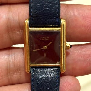 Könnte beinhalten: Eine rechteckige goldfarbene Armbanduhr mit dunkelburgunderfarbenem Zifferblatt, das den Schriftzug 'Must de Cartier' und ein goldenes Logo zeigt. Die Uhr hat dünne goldene Zeiger und ein dunkelblaues, strukturiertes Lederarmband mit blauem Cabochon.