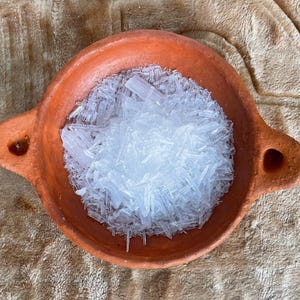 May include: A small, round, terracotta-coloured bowl with two handles, filled with a pile of white, crystalline structures. The bowl sits on a textured, tan surface, creating a contrast between the earthy tones and the bright crystals.