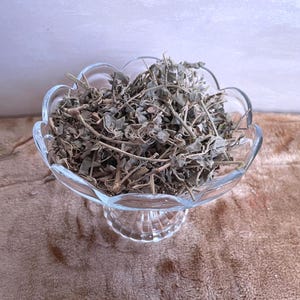 May include: A clear, scalloped-edge glass bowl filled with dried herbs. The bowl sits on a clear pedestal base, and the herbs are a mix of green and brown. The background is a soft, neutral color.