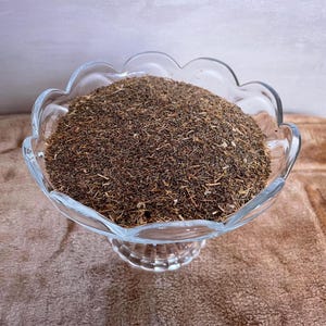 May include: A clear glass bowl with a decorative, scalloped edge, filled with dried, brown herbs. The bowl is on a brown surface, with a neutral background. The herbs appear finely chopped and have a slightly textured appearance.