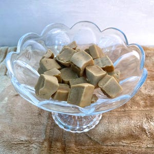 May include: A clear glass pedestal bowl with a scalloped rim filled with numerous small, light brown, cube-shaped pieces of fudge. The glass bowl features a textured, fluted base and a smooth, transparent body that holds the dense, tan-coloured confection.