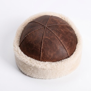 May include: A brown leather skullcap featuring a distressed texture and a six-panel construction with visible stitching. The hat is finished with a thick, cream-colored shearling trim that encircles the base, providing a contrasting border.