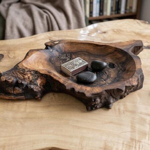 Pode incluir: Uma tigela de madeira artesanal com uma forma natural e orgânica. A tigela contém duas pedras pretas lisas e uma pequena caixa de prata ornamentada. Um pequeno objeto de latão está à direita. A tigela está sobre uma mesa de madeira clara.
