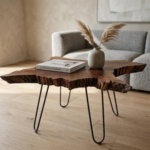 May include: A live-edge coffee table with a natural wood grain finish. The table has black hairpin legs and holds a book titled "CURATED FORMS" and a small vase with dried grasses. The table is approximately 76 cm long.