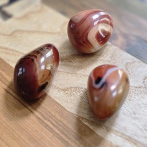 May include: Three polished agate stones in shades of red, brown, and cream, with unique swirling patterns. The stones are smooth and egg-shaped, displayed on a wooden surface.