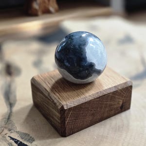 May include: A decorative sphere with a marbled pattern in dark blue, grey, and white, displayed on a small, rectangular wooden block. The wooden block has visible grain and is approximately 5 cm wide. The sphere has a polished, reflective surface.