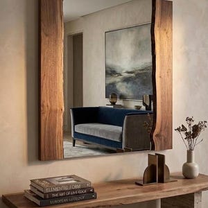 May include: A rectangular mirror with a natural wood frame. The frame has a live edge design. The mirror reflects a room with a blue sofa, artwork, and a wooden console table. Books and decorative items are on the table.