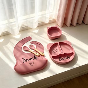 May include: A dusty pink silicone baby feeding set including a bib, a three-section bear-shaped plate, a small bowl, a spoon, and a fork. The bib and plate are personalized with the name Beverly in black script and a small heart icon.