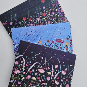 以下が含まれることがあります： 3つの花の絵が描かれたアートプリント。一番上のプリントは、暗い背景に赤い花と紫の花が描かれています。真ん中のプリントは、赤い花と黄色の花が描かれた青い背景です。一番下のプリントは、暗い背景にピンクの花が描かれています。