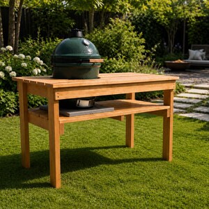 May include: A rectangular wooden grill table with four legs and a lower shelf, featuring a dark green ceramic kamado-style grill with a silver temperature gauge and a wooden handle mounted on the top surface of the table.
