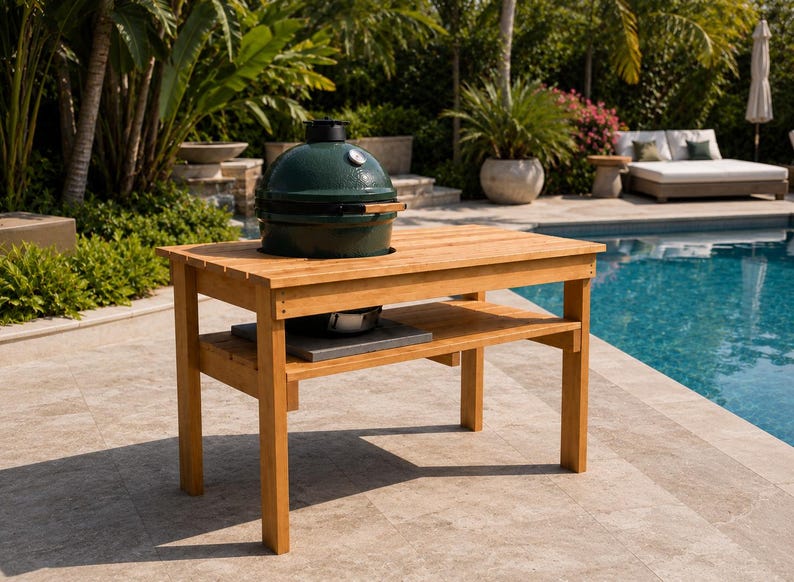 May include: A rectangular wooden grill table with a light brown finish, featuring a lower shelf and a circular cutout in the top surface. A dark green ceramic kamado-style grill with a silver temperature gauge is set into the table top.