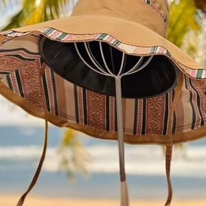 May include: A wide-brimmed sun hat in a tan color, featuring a patterned band around the crown and brim. The underside of the brim has a striped and patterned fabric. The hat has long, tan ties. The hat is displayed on a white wire stand.