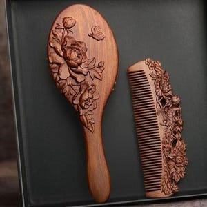 May include: A wooden hairbrush and comb set. The brush is oval-shaped with a handle, and the comb is rectangular. Both are intricately carved with floral and butterfly designs. The wood has a warm, reddish-brown tone.
