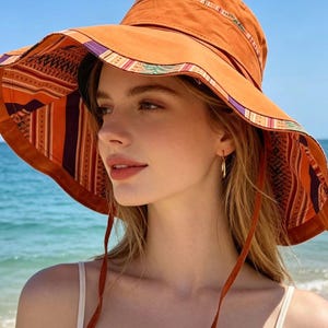 May include: An orange sun hat with a wide brim, featuring a patterned interior and trim with purple, green, and white accents. The hat has a chin strap and is worn outdoors near the ocean.