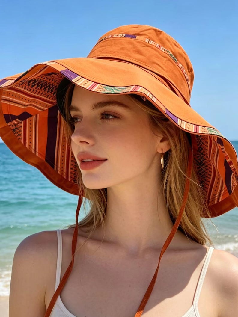 May include: An orange sun hat with a wide brim, featuring a patterned interior and trim with purple, green, and white accents. The hat has a chin strap and is worn outdoors near the ocean.