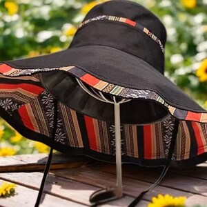 May include: Black sun hat with a wide brim and a patterned band. The brim's underside features a colorful geometric design with red, orange, and black stripes. A black drawstring is attached. The hat is displayed on a white stand against a blurred background of sunflowers.