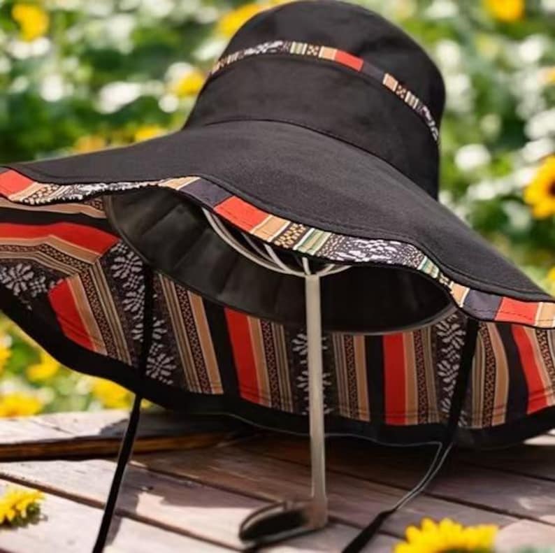 May include: Black sun hat with a wide brim and a patterned band. The brim's underside features a colorful geometric design with red, orange, and black stripes. A black drawstring is attached. The hat is displayed on a white stand against a blurred background of sunflowers.