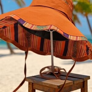 May include: A wide-brimmed sun hat in a warm orange hue, featuring a patterned trim in shades of orange, purple, and green. The interior lining showcases a vibrant striped design. The hat is displayed on a small wooden stool, with brown straps hanging down.
