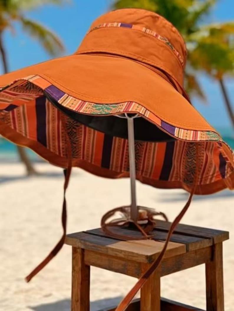 May include: A wide-brimmed sun hat in a warm orange hue, featuring a patterned trim in shades of orange, purple, and green. The interior lining showcases a vibrant striped design. The hat is displayed on a small wooden stool, with brown straps hanging down.