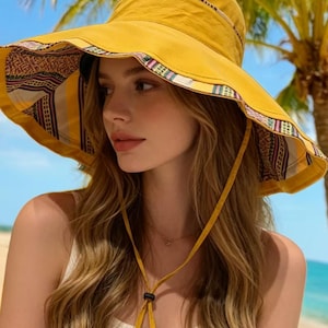 May include: A yellow sun hat with a wide, wavy brim. The interior brim features a patterned fabric with red, brown, and cream colors. The hat has a chin strap and is worn by a person with long, wavy hair. The background includes a beach and palm trees.