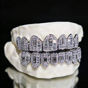 Può includere: Un paio di grillz incastonate di diamanti, esposte su un calco dentale bianco. Le grillz presentano diamanti taglio rettangolare, creando un effetto scintillante. Lo sfondo è nero, evidenziando i gioielli.
