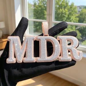 Op de afbeelding: Een roségouden hanger met de letters "MDR" met een zilver en diamantachtig ingelegd ontwerp. De hanger wordt vastgehouden door een gehandschoende hand voor een raam met uitzicht op bomen.