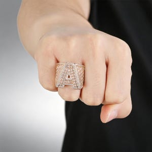 Puede incluir: Un anillo en tono oro rosa que presenta una letra A grande de estilo bloque en la parte frontal. Toda la superficie de la letra y la banda ancha están incrustadas con numerosas piedras pequeñas, claras y brillantes en un engaste pavé.
