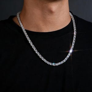 Può includere: Una collana a catena color argento caratterizzata da una fila continua di pietre rettangolari, trasparenti e sfaccettate, incastonate in una montatura metallica. La collana poggia su uno sfondo di tessuto nero con finitura lucida.