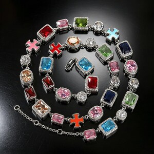 May include: A silver-toned necklace featuring a variety of colorful gemstones in square, oval, and round shapes. The gemstones are in shades of red, pink, blue, green, and orange, with a few clear crystals. The necklace has a lobster clasp.