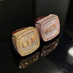 May include: Two square-faced rings with a gold and rose gold finish. Each ring is encrusted with small, sparkling stones. One ring has the letters "LET" and the other has "CRY" in the center.