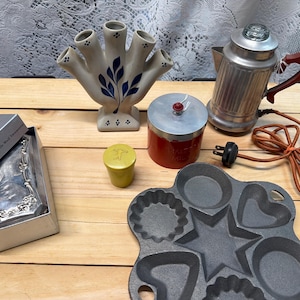 May include: A selection of vintage kitchenware on a wooden surface. Includes a silver coffee pot, a red and silver container, a cast iron muffin pan, a ceramic vase, a silver serving piece, and a yellow container.