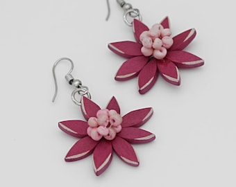Peut inclure: Une paire de boucles d'oreilles en forme de fleurs rose et bordeaux avec des crochets argentés. Les fleurs sont en bois et ont un centre rose.