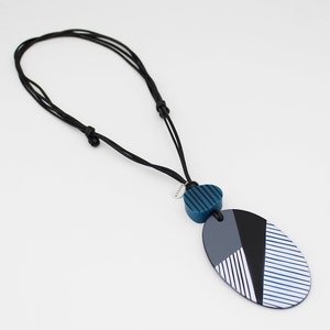 May include: A modern necklace with a black cord and a geometric pendant. The pendant features a combination of blue, gray, black, and white shapes. The necklace has a small, round tag with text.