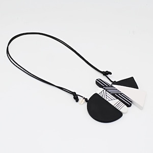 May include: A geometric necklace in black and white, suspended from a black cord. The pendant comprises a black semi-circle, a white triangle, and a striped element. The necklace is set against a white backdrop.