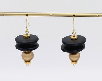 May include: A pair of dangle earrings featuring a gold-colored hook and accents. Each earring has two stacked black, round discs above a gold-colored sphere. The earrings hang from a gold bar.