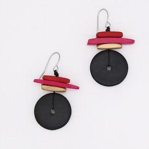 May include: A pair of handcrafted earrings featuring a black circular base with a small hole. Above the base are stacked wooden pieces in red, pink, and gold. The earrings have silver-colored ear wires and a black string connecting the wooden elements.
