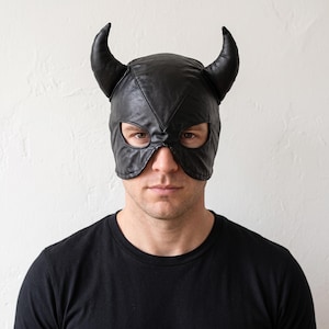 May include: A black leather mask featuring two prominent, curved horns extending from the top. The mask covers the upper portion of the face, including the forehead and temples, with two distinct eye openings and a V-shaped seam across the bridge.
