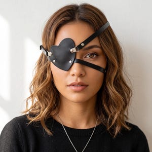 May include: A black heart-shaped eye patch made of smooth material, featuring thin adjustable straps that secure around the head with silver-toned metal hardware and rivets. The patch covers the right eye, held in place by a strap across the forehead.