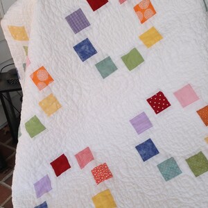 K&ouml;nnte beinhalten: Eine wei&szlig;e Steppdecke mit Patchwork-Muster, das aus Quadraten in verschiedenen Farben besteht, darunter Rot, Gelb, Gr&uuml;n, Blau und Lila.