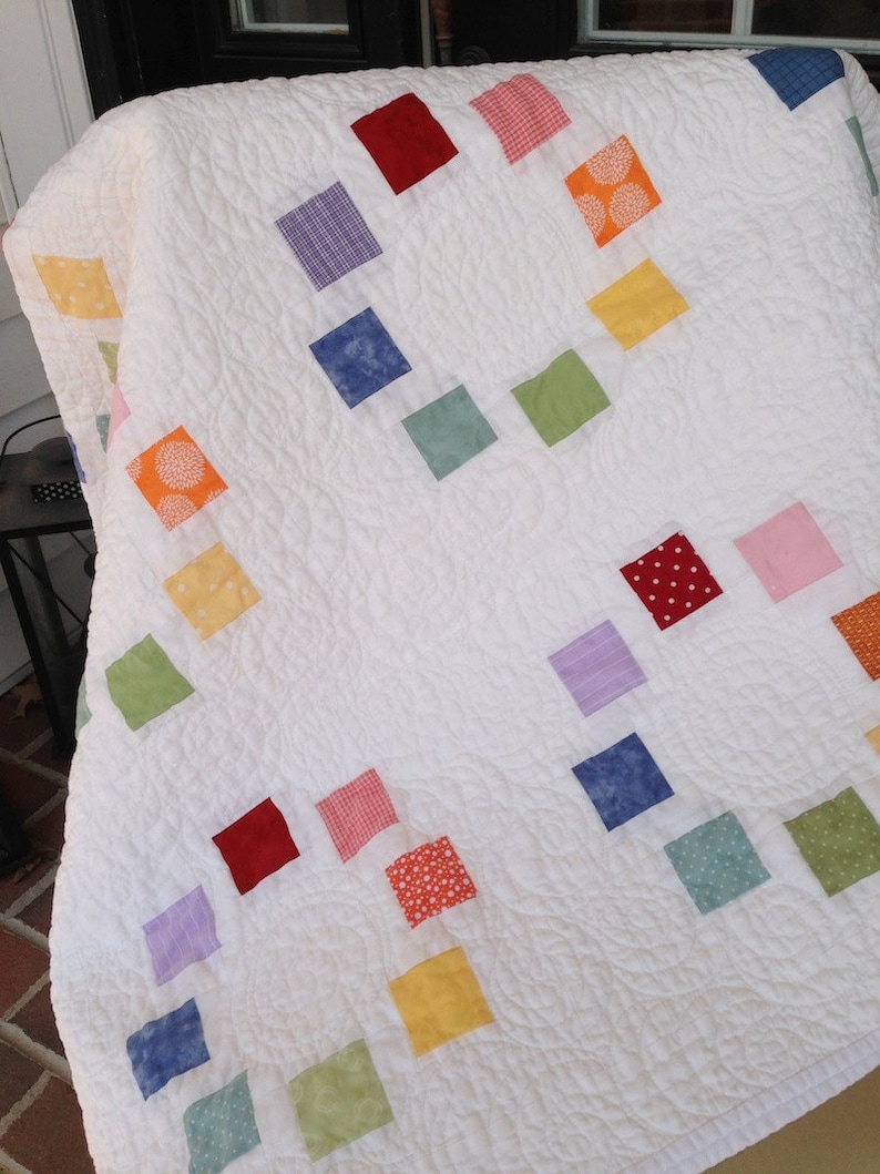 K&ouml;nnte beinhalten: Eine wei&szlig;e Steppdecke mit Patchwork-Muster, das aus Quadraten in verschiedenen Farben besteht, darunter Rot, Gelb, Gr&uuml;n, Blau und Lila.